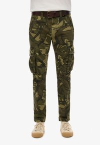 Cargo pants in een groene camouflageprint, gemaakt van duurzaam materiaal, hebben zijzakken en een slanke pasvorm. Draag ze met een bruine riem en lichte sneakers.