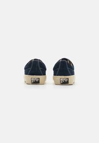 Sapatos de camurça azul-marinho com bico redondo, sola de borracha bege, costura branca e um logótipo no calcanhar. Design minimalista sem atacadores.
