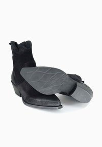 Un paio di stivali neri in suede alla caviglia con punte appuntite, tacchi a blocco e suole in gomma texturizzata su uno sfondo bianco.