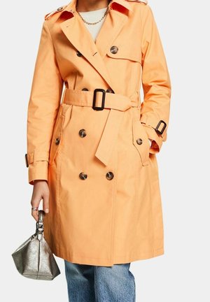 Person iført en ferskenfarvet dobbeltradet trenchcoat med bælte, der holder en lille metallisk håndtaske, kombineret med blå jeans og en guldkæde rundt om halsen.