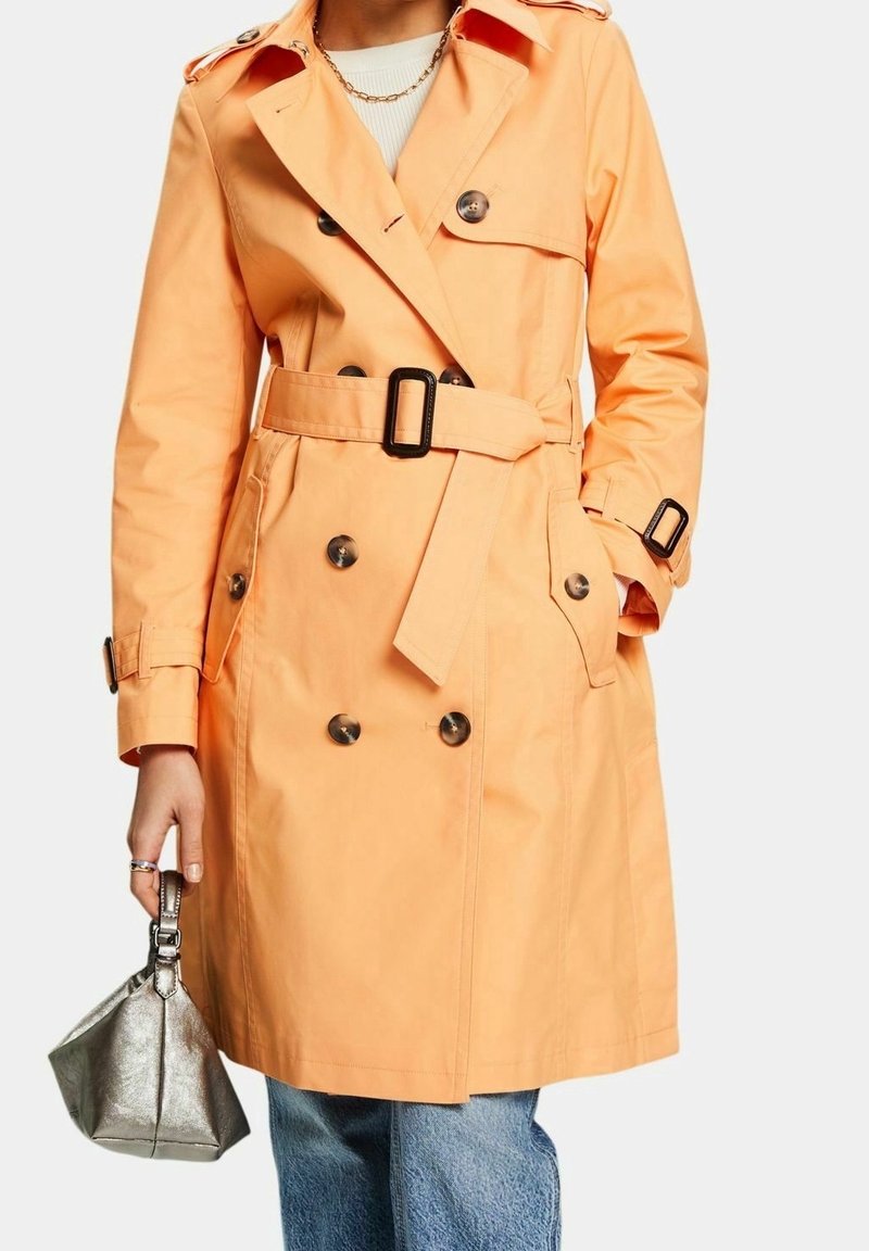 Personne portant un trench-coat double boutonnage pêche avec ceinture, tenant un petit sac à main métallique, associé à un jean bleu et un collier chaîne doré.