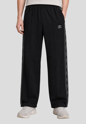 Pantaloni da ginnastica neri con strisce laterali a motivi a diamante, vita elasticizzata e piccolo logo Umbro sulla coscia sinistra, indossati con sneakers bianche.