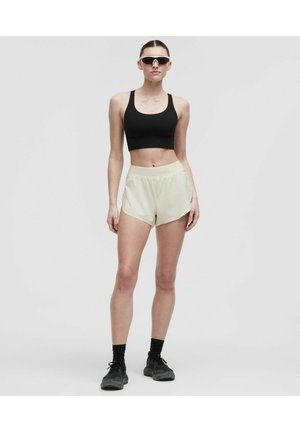 Woman wearing black sports bra, light shorts, black socks, black sneakers, and white sports sunglasses standing against a plain gray background.