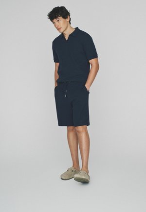 Młody mężczyzna w granatowej polo z krótkim rękawem i pasujących szortach oraz beżowych wsuwanych butach, stojący z rękami w kieszeniach.