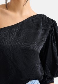 Schwarzes Top mit einem Schulterträger aus strukturiertem Stoff, der dezente Wellenmuster und dramatische Puffärmel aufweist, ergänzt durch einen lockeren Schnitt.