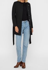 Black wrap coat with a minimalistic design and silver buttons, paired with light blue jeans and brown heeled boots.