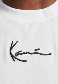 White t-shirt featuring a black embroidered signature design. Fabric appears soft with a smooth texture and crew neckline.