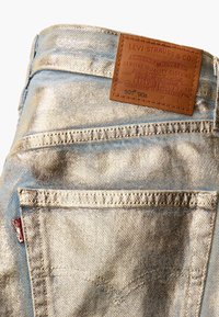 Jeans in denim blu chiaro con finitura metallica. Presentano una toppa in pelle marrone con dettagli in rilievo e tasche posteriori con accenti di cucitura.
