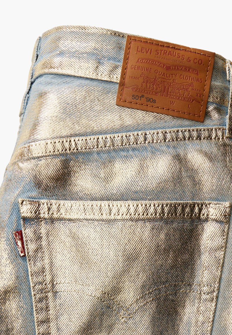 Jean en denim bleu clair avec un éclat métallique. Comprend une patch en cuir marron avec des détails en relief et des poches arrière avec des accents de couture.
