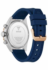Arrière en acier inoxydable argenté d'une montre Guess avec un bracelet en silicone bleu et une boucle dorée, affichant des informations sur la résistance à l'eau et le modèle.