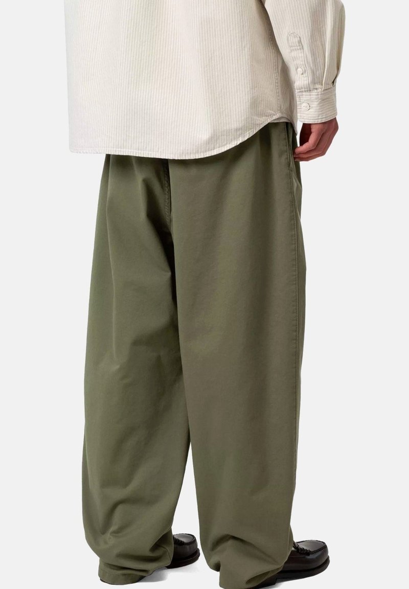 Persona vestida con pantalones sueltos de color verde oliva, una camisa beige de manga larga y zapatos negros, de pie con la mano derecha visible.