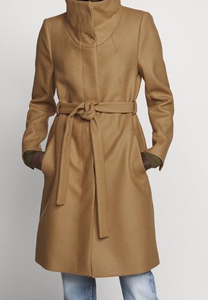Long manteau en laine marron camel avec col montant et ceinture à nouer, porté sur un jean bleu avec les mains dans les poches latérales.