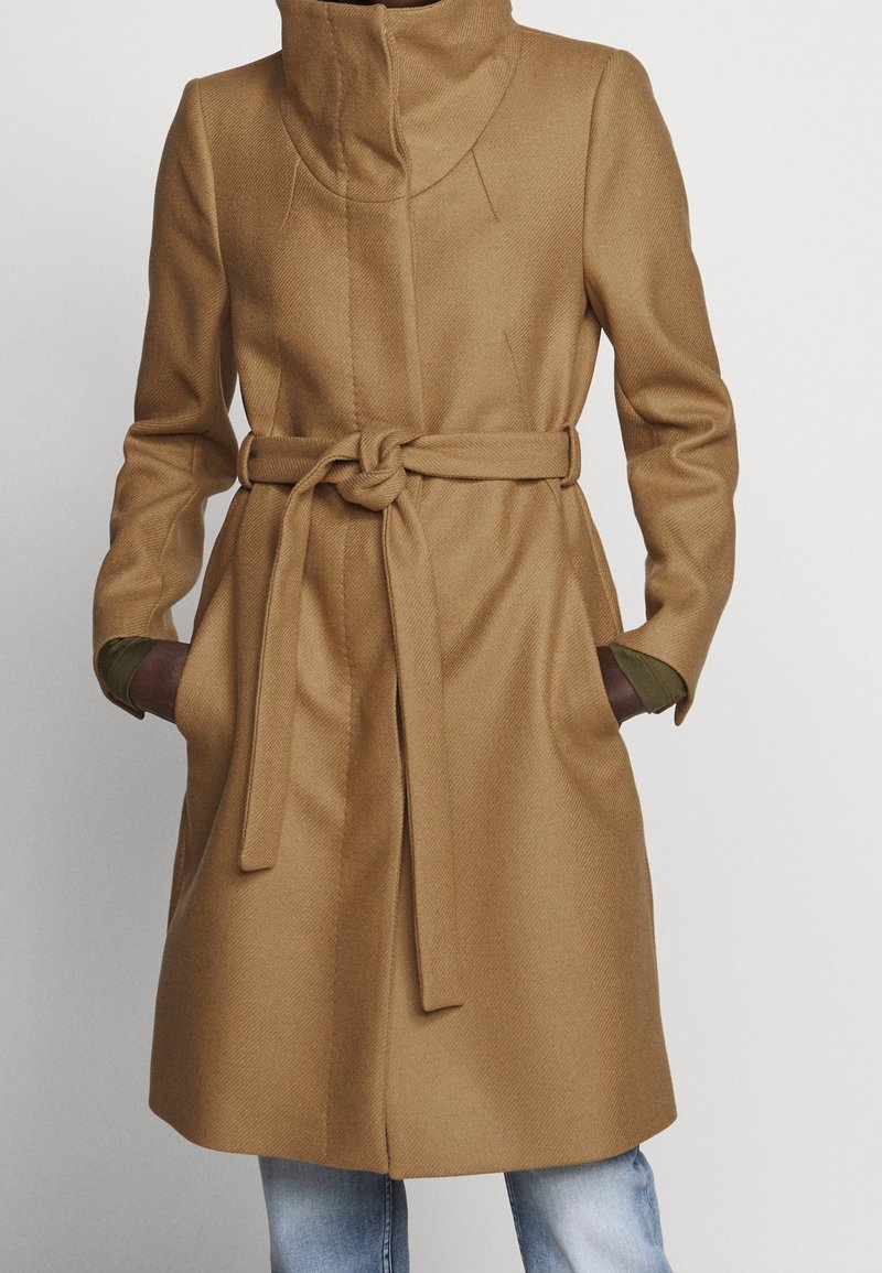 Long manteau en laine marron camel avec col montant et ceinture à nouer, porté sur un jean bleu avec les mains dans les poches latérales.
