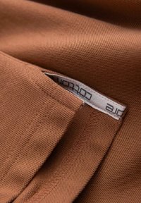 Brun bomullstyg med en texturerad väv, med en sydd etikett synlig inom en vikt kant. Enkel konstruktion utan utsmyckningar.