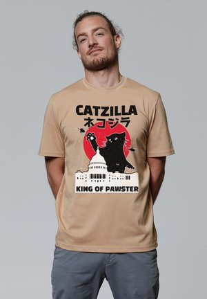 Beige t-shirt met korte mouwen met een zwart grafisch ontwerp van een kat, tekst en een silhouet van een gebouw, met een rode cirkelvormige accent achter de kat.