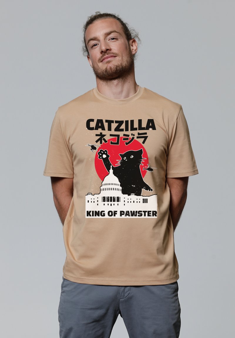 Beige t-shirt met korte mouwen met een zwart grafisch ontwerp van een kat, tekst en een silhouet van een gebouw, met een rode cirkelvormige accent achter de kat.
