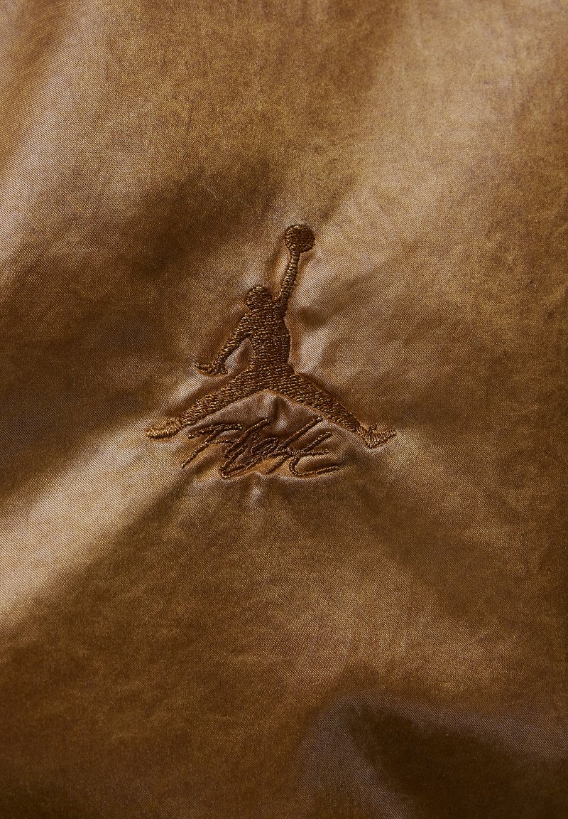 Braunes Satin-Gewebe mit einem gestickten Jumpman-Logo und dem Wort "Flight" darunter in einer Schreibschrift.