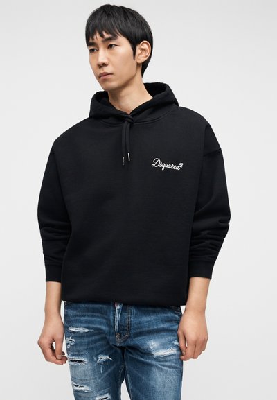 Μαύρο φούτερ με κουκούλα, εμπρόσθια τσέπη, κορδόνι για κλείσιμο και λευκό λογότυπο. Φορεμένο με σκισμένο μπλε τζιν, παρουσιάζοντας ένα καθημερινό στυλ.