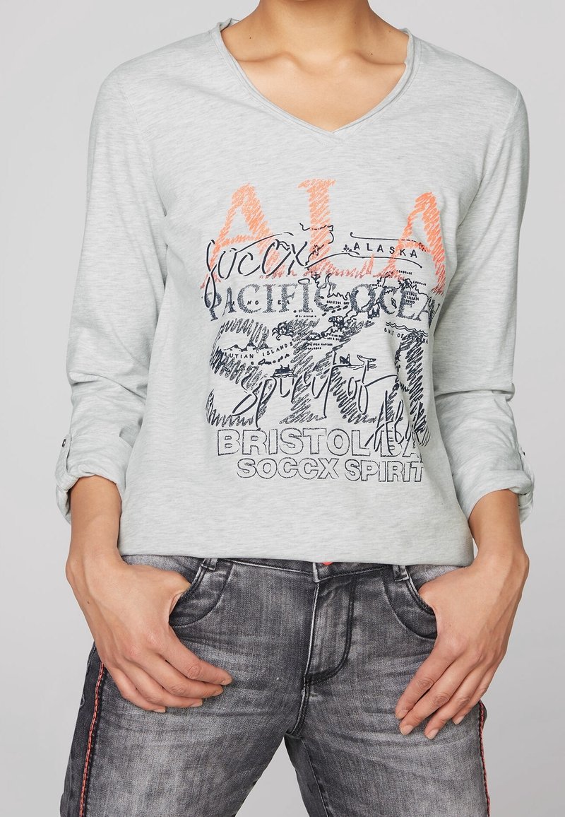 Světle šedá dlouhá košile s krátkým rukávem, s výstřihem do V, s grafikou a textem v černé a oranžové barvě. Měkký materiál s ohrnutými manžetami.
