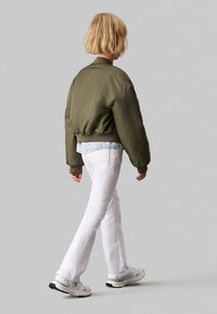 Groene bomberjack met geribbelde manchetten en zoom, gelaagd over een licht spijkerhemd, gecombineerd met witte flared jeans en witte sneakers.