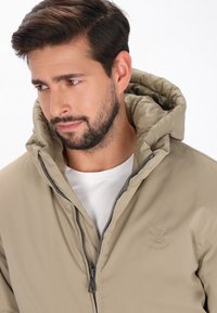 Beige polstret jakke med hætte, med lynlås lukning, tekstil tekstur og et præget ankerlogo på brystet. Hvid skjorte under.