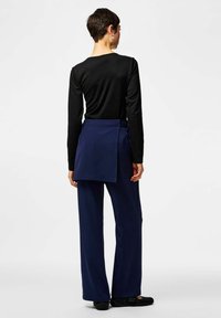 Chemise noire à manches longues associée à un pantalon large bleu marine et un tablier assorti à nouer sur le côté.