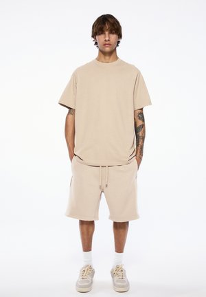 Junger Mann mit Tattoos, der ein beiges oversized T-Shirt, passende Shorts, weiße Socken und Sneakers trägt und vor einem schlichten Hintergrund steht.