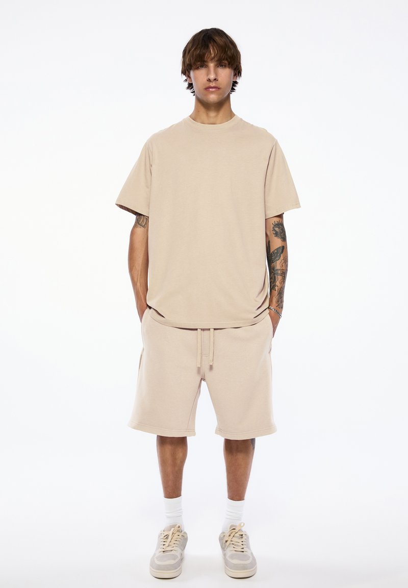 Jeune homme tatoué portant un t-shirt beige oversize, un short assorti, des chaussettes blanches et des baskets, debout devant un fond uni.