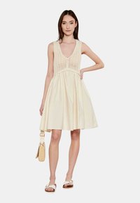 Vestido de sol color crema con un cuerpo de ganchillo, falda acampanada y escote en V. Usado con sandalias planas y sosteniendo un pequeño bolso de mimbre.