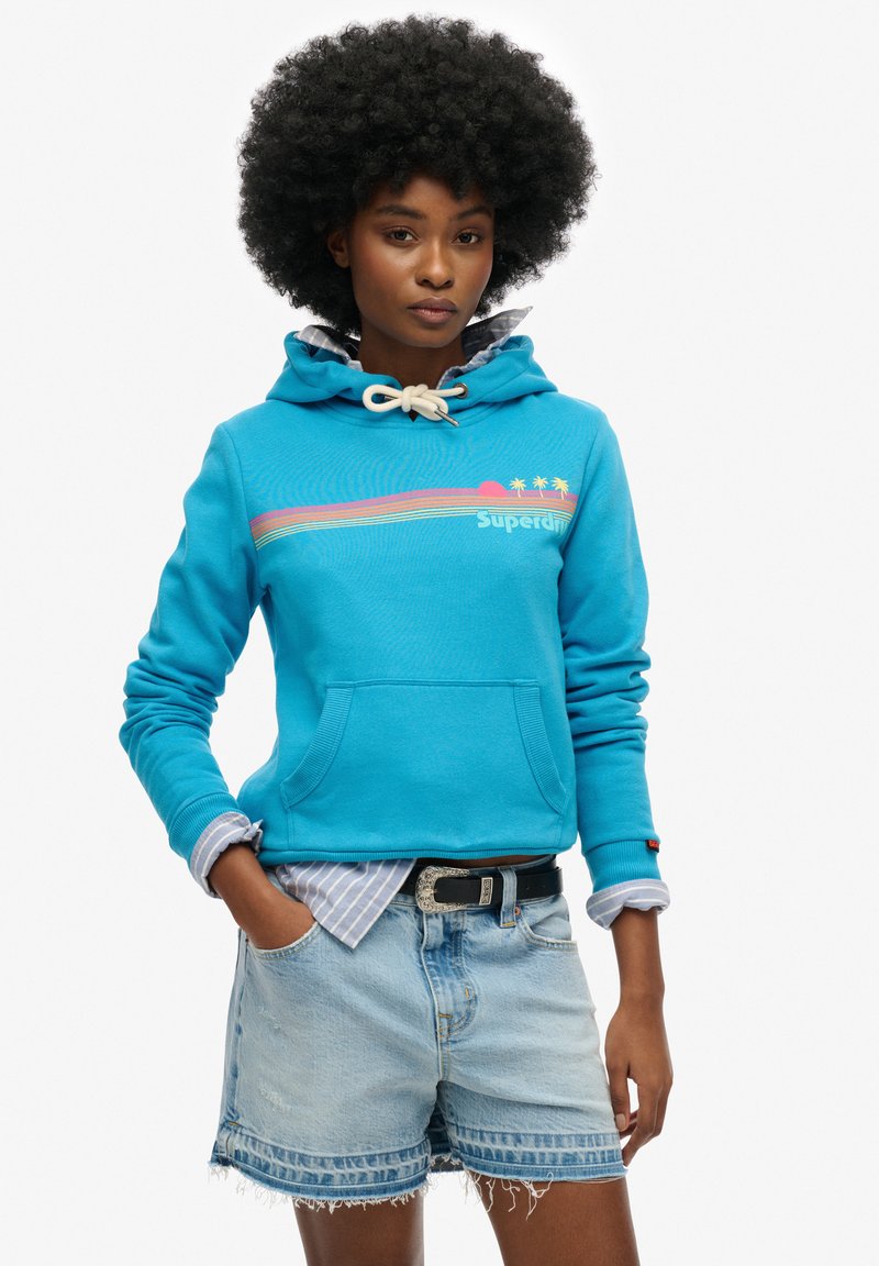 Blauwe hoodie met een kangoeroezak en koord. Voorzien van kleurrijke grafische strepen en het "Superdry" logo. Gepaard met denim shorts.