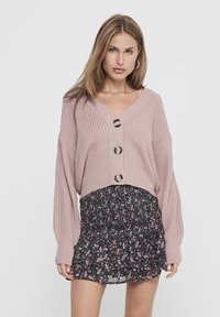 Lichtroze, gebreide cardigan met een V-hals, oversized mouwen en drie ronde knopen. Gecombineerd met een zwarte rok met bloemenprint.