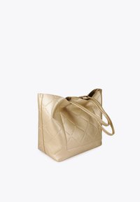 Złota torebka z pikowanego materiału z długimi uchwytami, ustawiona pod kątem na jasnym tle. Materiał ma błyszczące wykończenie.