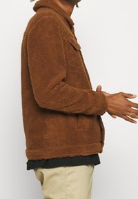 Chaqueta de forro polar marrón con una superficie texturada, que presenta un cuello, bolsillos laterales y un ajuste holgado. Llevada sobre una camisa negra.