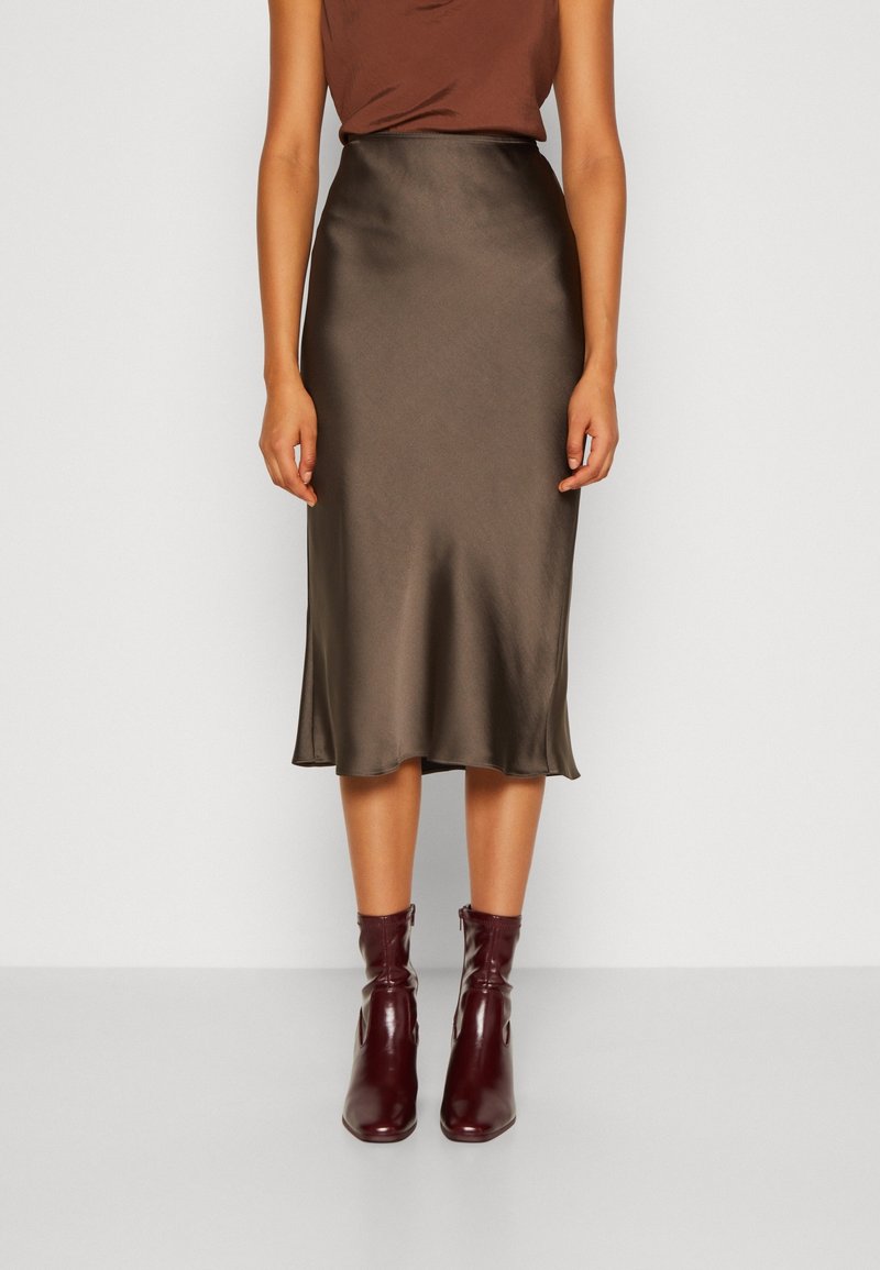 Femme portant une jupe midi en satin marron et des bottines bordeaux, debout devant un fond blanc uni.