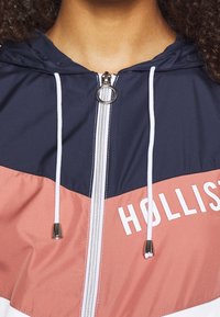 Huvtröja med vindskydd i marinblått och korallfärgade paneler. Har en silverfärgad dragkedja, vita snoddar och en tryckt logotyp i vitt på bröstet.