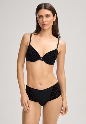 Woman standing facing forward wearing matching black textured bra and high-waisted panties against plain light gray background.