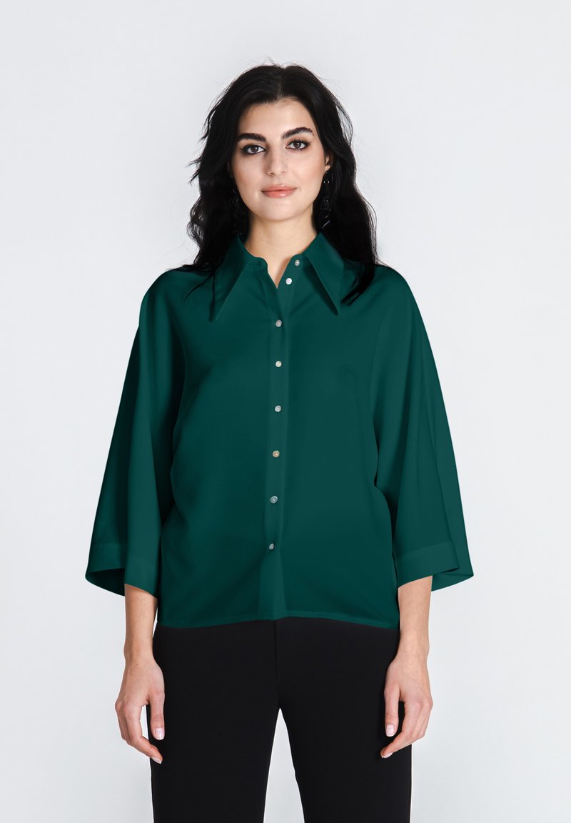 Türkise lahtiseid blouse, millel on terav krae, hõbedased nupud ja 3/4 varrukad, koos mustade pükstega. Sile kangatekstuur.