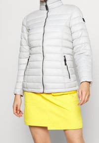 Giacca imbottita grigio chiaro con motivo trapuntato verticale, colletto alto, cerniera frontale e tasche con zip nere. Indossata sopra una gonna gialla.