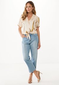 Korte mouw blouse in roomkleur met gele en zwarte abstracte patronen, vastgebonden in de taille; gecombineerd met lichtblauwe high-waisted jeans en gouden hakken.