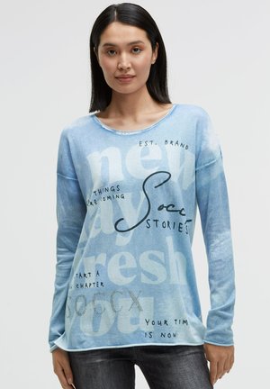 Frau mit glatten schwarzen Haaren, die ein hellblaues Langarmshirt mit gemischtem Text trägt, darunter "new", "fresh" und "Soccx", sowie eine dunkle Hose.