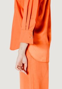 Blusa arancione brillante con una texture setosa, caratterizzata da un orlo arcuato, polsini abbottonati e pieghe sottili lungo la manica.