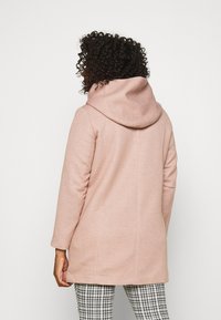 Rosa huva med en strukturerad finish, skräddarsydd passform och långa ärmar. Har en mjuk, draperad huva. Bärs över byxor med rutigt mönster.