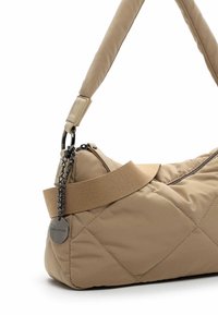 Sac en bandoulière matelassé beige avec une sangle rembourrée, une fermeture éclair et un détail en chaîne argentée. Il présente une étiquette en métal de la marque et un design épuré et minimaliste.
