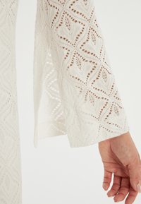 Witte stof met een diamantpatroon, met een gestructureerde uitstraling en uitlopende mouwen. Hand die naar de rand van de mouw reikt.
