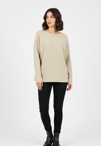 Frau mit schulterlangem, welligem Haar, die einen beigen, strukturierten Pullover, schwarze enge Hosen und schwarze Stiefeletten trägt und vor einem weißen Hintergrund steht.