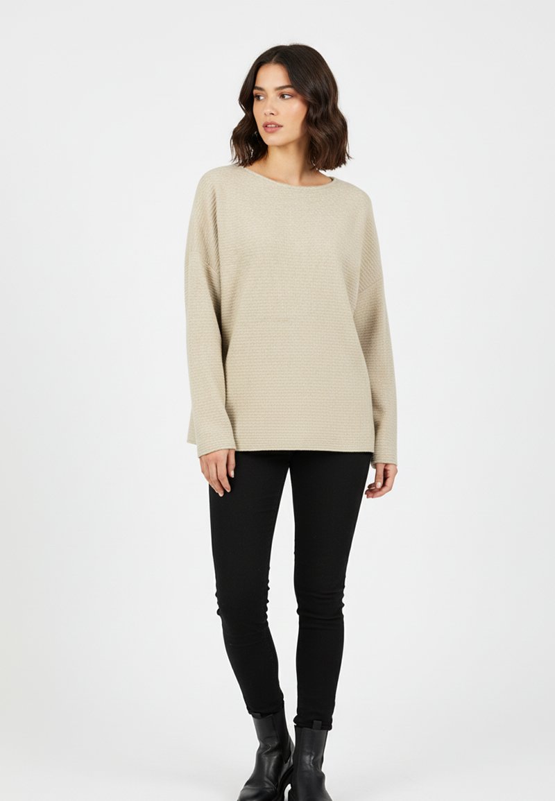 Frau mit schulterlangem, welligem Haar, die einen beigen, strukturierten Pullover, schwarze enge Hosen und schwarze Stiefeletten trägt und vor einem weißen Hintergrund steht.