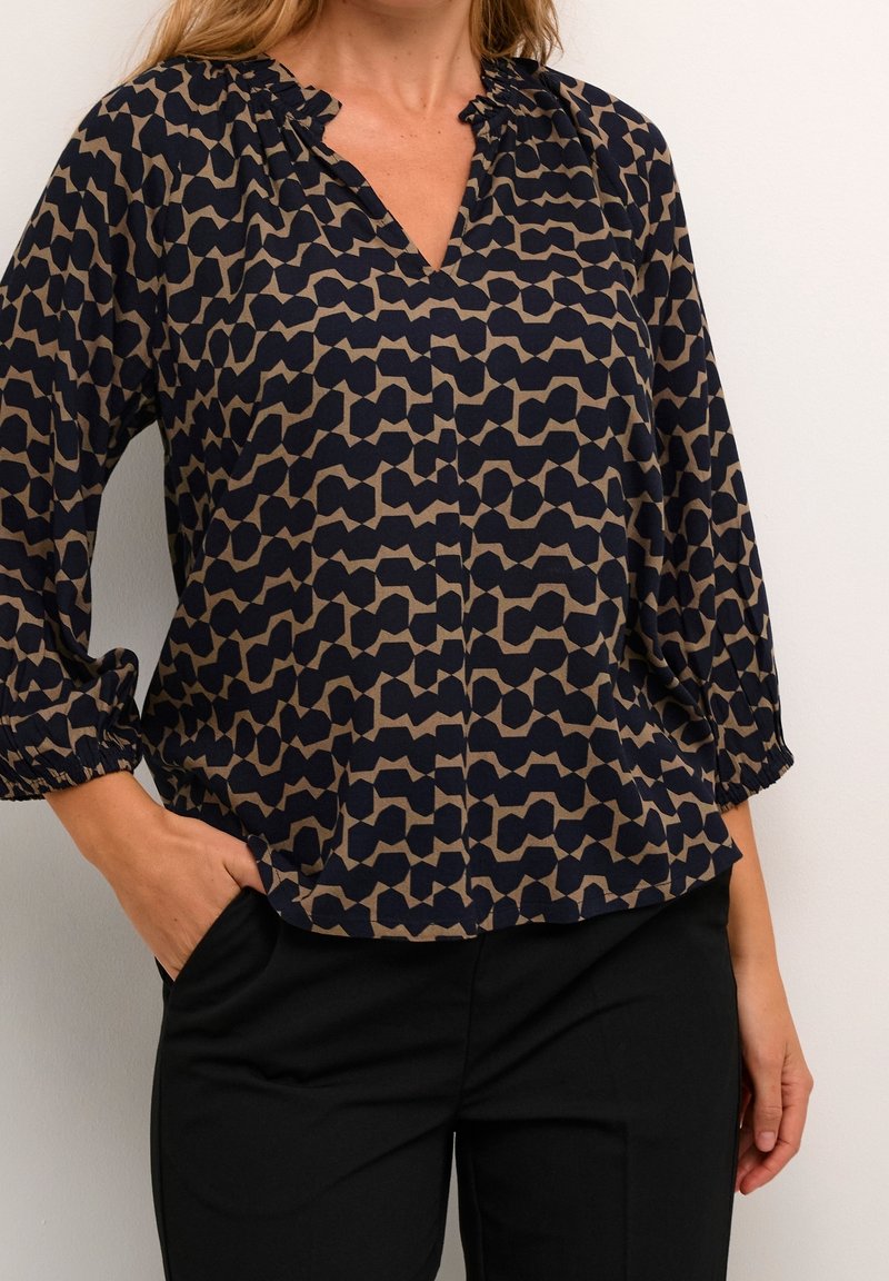 Blouse marine à motif abstrait beige, col en V et poignets élastiques. Tissu doux qui tombe doucement, avec une coupe décontractée et un léger éclat.
