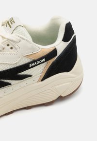 Sneaker featuring a cream mesh upper with beige and black suede accents, wave pattern design, and a textured off-white midsole.
