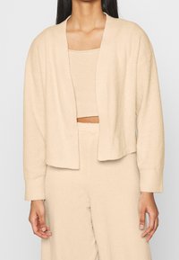 Cardigan lavorato a maglia beige con apertura frontale, polsini a coste e tessuto morbido e testurizzato. Indossato sopra un top coordinato.