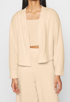 Cardigan en tricot beige à l'avant ouvert, avec des poignets côtelés et un tissu doux et texturé. Porté par-dessus un débardeur assorti.